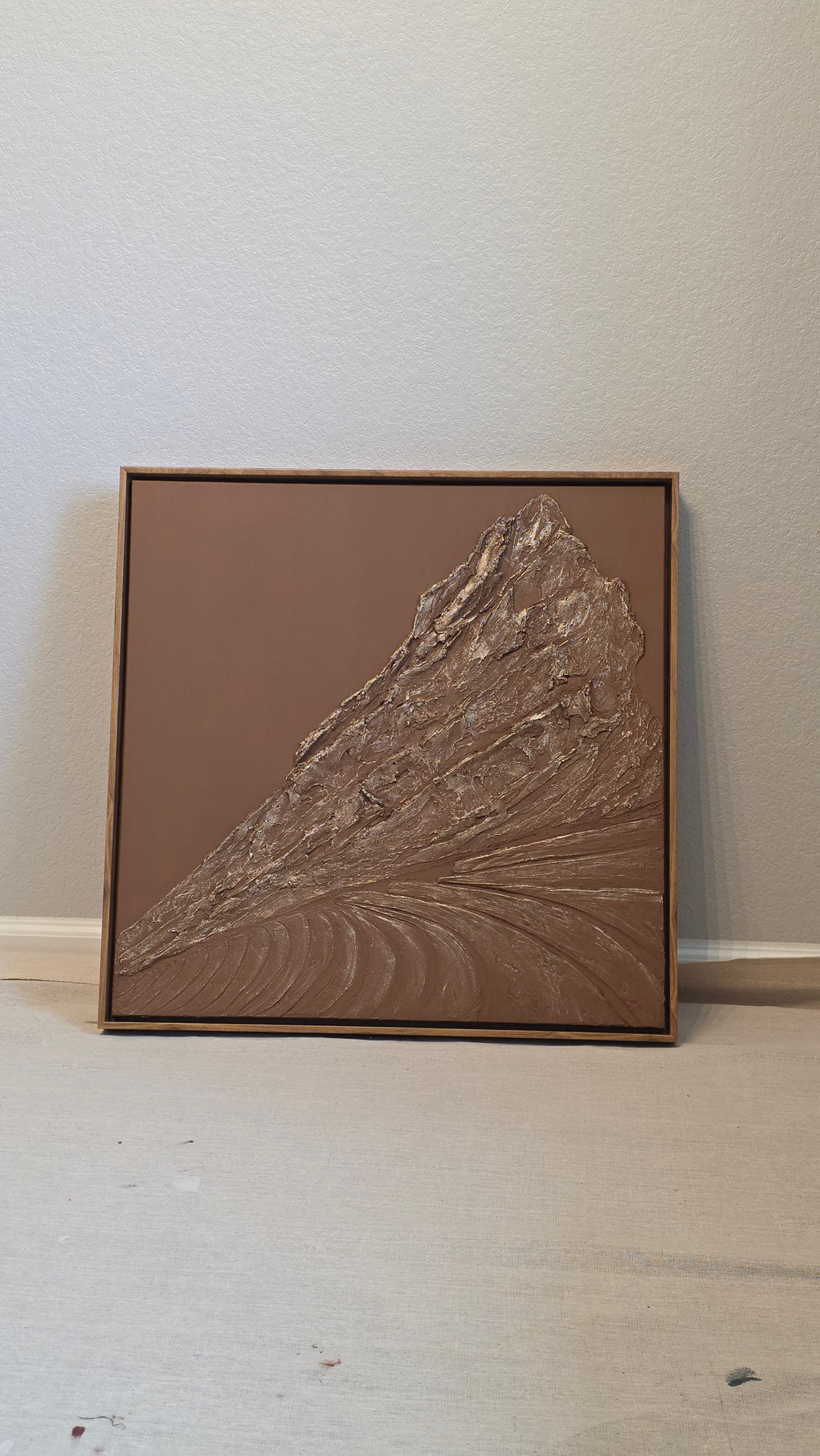 A square framed wall art piece featuring a textured, embossed fan or leaf-like design in metallic bronze and brown tones, mounted on a white wall.