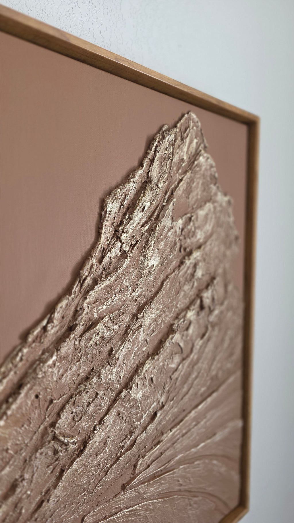 A square framed wall art piece featuring a textured abstract design with layered, ridged metallic-looking brown and silver forms on a warm taupe background.